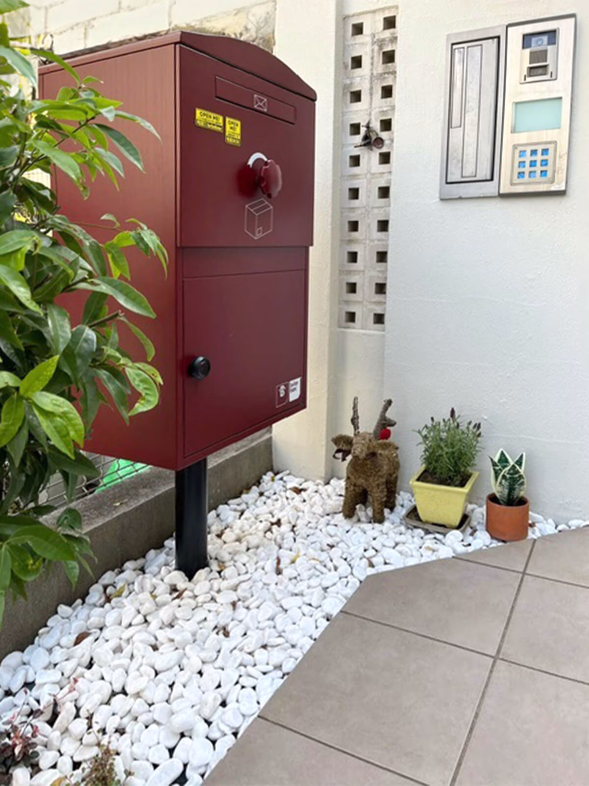 鉢植えの緑を添えた玄関脇に立つ、ボルドーの宅配ボックス